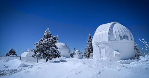 Snow-covered observatories surrounded by trees under a clear blue sky.