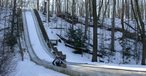 Two people sledding down a snowy hill, surrounded by trees and a wooden railing in a winter landscape.