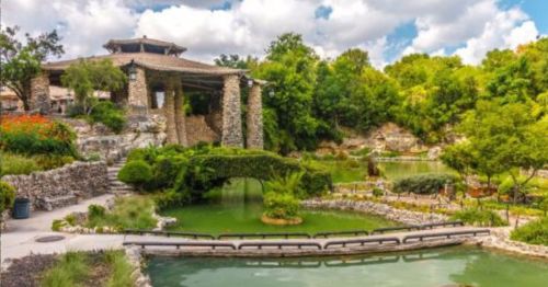 A serene garden with a pond, stone bridge, and lush greenery, featuring a pavilion in the background under a cloudy sky.