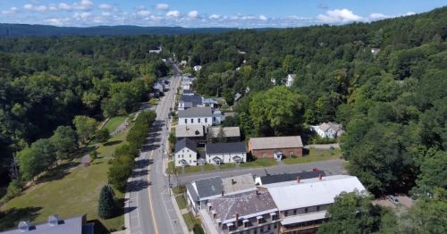 Aerial view of a small town surrounded by trees, featuring a main road and various buildings along the street.