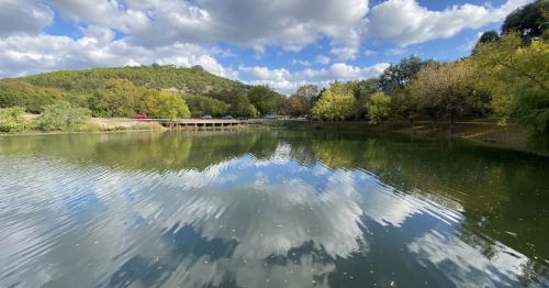 A serene lake reflecting clouds and trees, with a bridge and cars in the background under a partly cloudy sky.