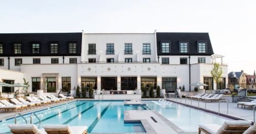 A modern hotel with a large outdoor pool surrounded by lounge chairs and greenery under a clear sky.