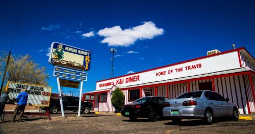 A diner with a sign reading "Home of the Travis," featuring cars parked outside under a blue sky with clouds.