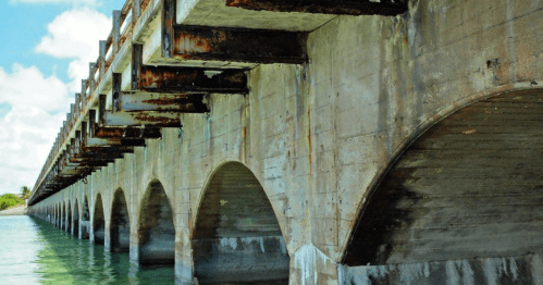 A weathered concrete bridge with arches, spanning over calm water under a blue sky with scattered clouds.