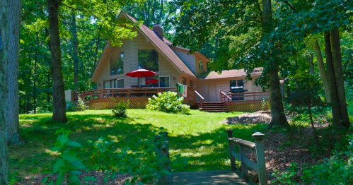 A charming house surrounded by trees, featuring a deck with a red umbrella and a lush green lawn.