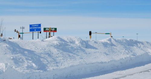 Snow-covered landscape with a tourist information sign and gas station visible in the background under a clear blue sky.