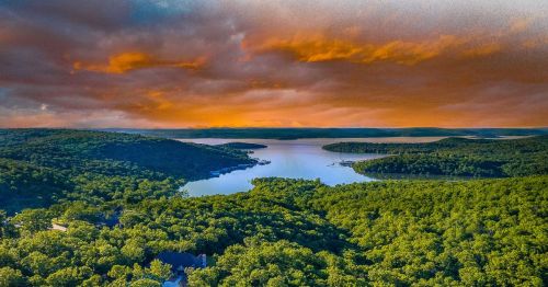 Aerial view of a serene lake surrounded by lush green forests under a dramatic orange and blue sky.