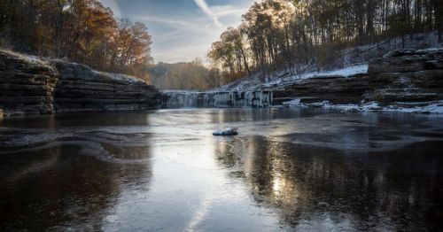 A serene winter landscape featuring a frozen river, rocky cliffs, and trees with autumn foliage under a clear sky.