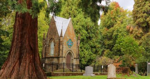 A small stone chapel surrounded by trees in a cemetery, with a decorative window and gravestones nearby.