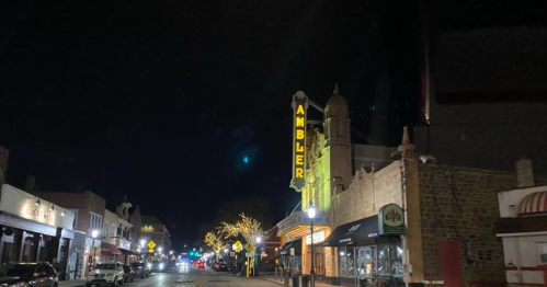 A nighttime view of a street with the Ambler Theater sign illuminated and festive lights on nearby buildings.