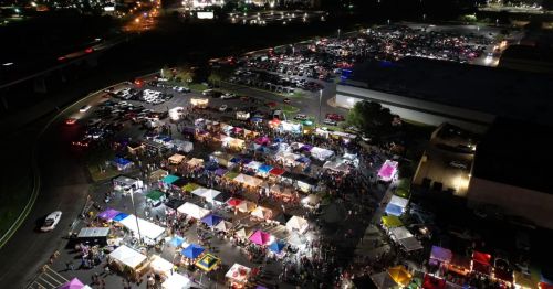 Aerial view of a vibrant night market with colorful tents and a large crowd, surrounded by parked cars and city lights.