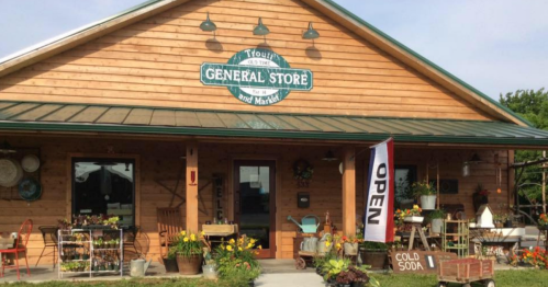 A rustic general store with a wooden facade, open sign, and colorful flowers in front.