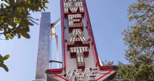 A vintage neon sign reading "MEAT HERE" with bright lights and an arrow, set against a clear blue sky.