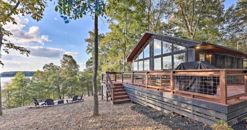 A modern cabin with large windows, surrounded by trees, overlooking a lake, with a deck and outdoor seating area.