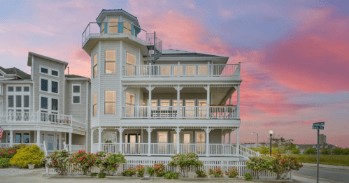 A large, multi-story beach house with balconies, surrounded by colorful flowers, under a vibrant sunset sky.
