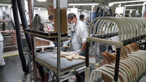 A worker in a factory carefully shapes wooden guitar bodies on a production line, surrounded by tools and equipment.