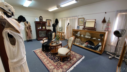 A vintage room displaying historical clothing, furniture, and artifacts, with a decorative rug and soft lighting.