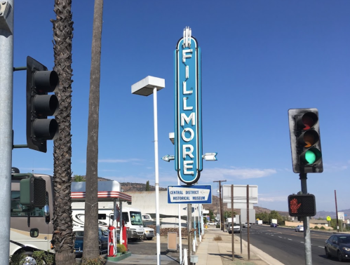 A bright blue "Fillmore" sign stands beside a street with traffic lights and palm trees under a clear blue sky.