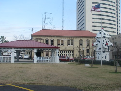 Historic building with a red roof, next to a large fire hydrant and a flagpole, set in a grassy area.