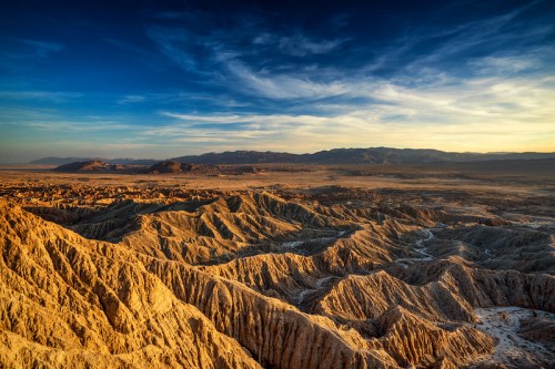 A vast desert landscape with rugged, textured hills under a blue sky and soft clouds at sunset.