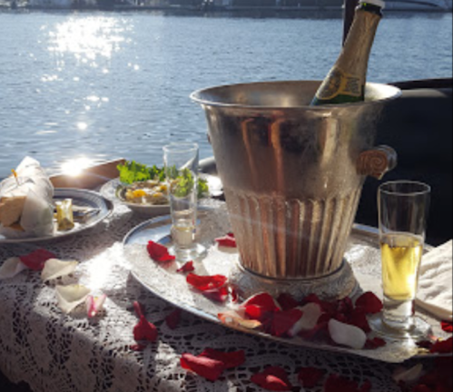 A romantic table setting by the water, featuring a champagne bucket, glasses, and rose petals scattered on the table.