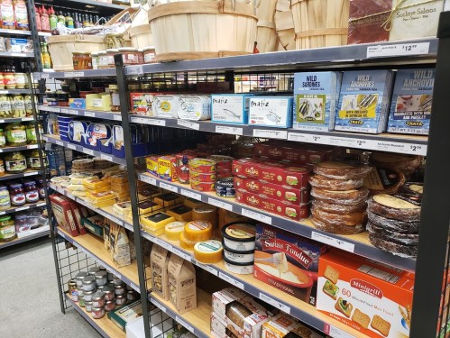 A grocery store aisle filled with various canned goods, cheeses, and packaged foods on shelves.