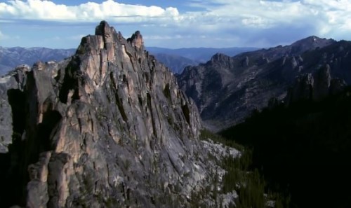 A rugged mountain landscape with steep rock formations and distant peaks under a partly cloudy sky.