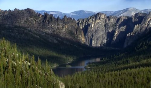 A scenic view of a river winding through a forested valley, surrounded by towering rocky cliffs and mountains.