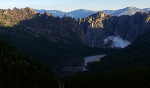 A scenic view of mountains and a lake, with sunlight casting shadows over the rugged terrain and surrounding forest.