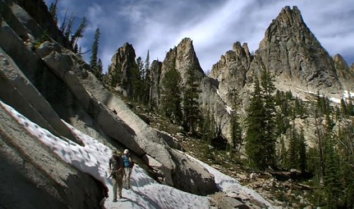 A hiker traverses a rocky, snowy landscape surrounded by tall, rugged mountains and pine trees under a cloudy sky.