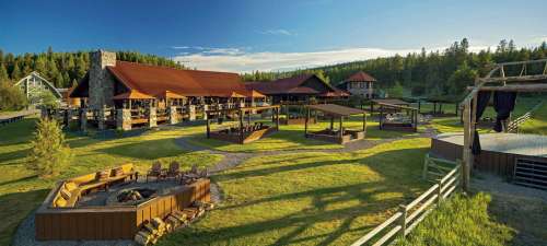 A spacious lodge with a wooden deck, surrounded by green grass and trees, featuring outdoor seating and a stage area.