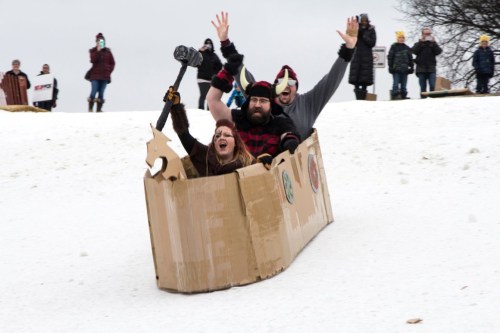 Three people joyfully sliding down a snowy hill in a cardboard sled, with onlookers cheering in the background.