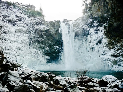 A frozen waterfall cascades into a turquoise pool, surrounded by snow-covered rocks and a misty, wintry landscape.