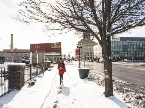 A woman walks on a snowy sidewalk, with trees and buildings lining the street on a sunny winter day.