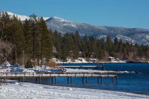 A snowy lakeside scene with a wooden pier, surrounded by evergreen trees and mountains in the background.