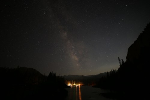 A serene night sky filled with stars and the Milky Way, reflecting over a calm lake surrounded by trees.