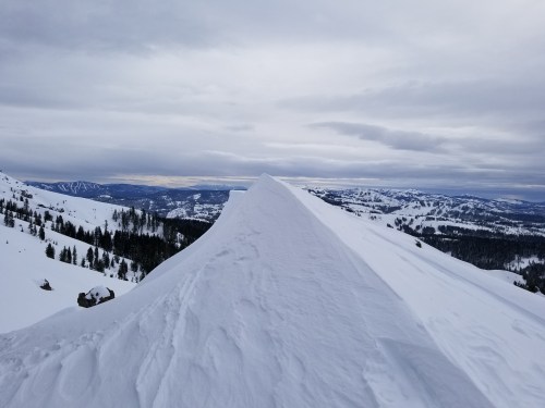 A snowy mountain peak with a ridge, surrounded by a vast winter landscape under a cloudy sky.