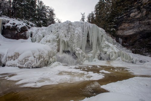 A frozen waterfall surrounded by snow and ice, with a river flowing in front, set in a wooded landscape.