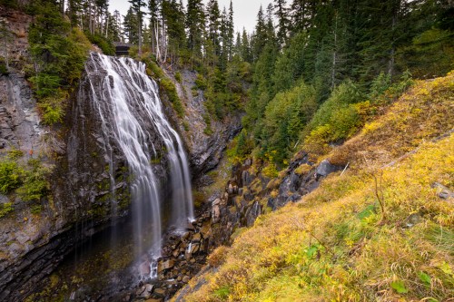 A serene waterfall cascades down rocky cliffs, surrounded by lush greenery and autumn foliage.