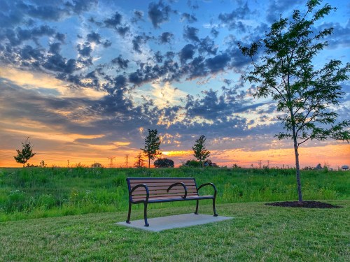 A park bench sits on a grassy area, framed by a vibrant sunset and scattered clouds in the sky.