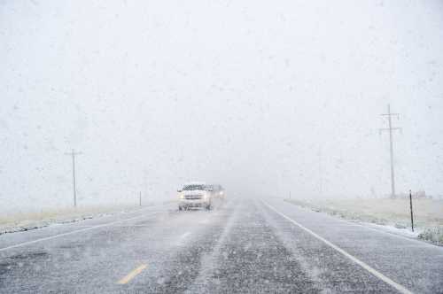 A snowy road scene with limited visibility, featuring vehicles driving through heavy snowfall.