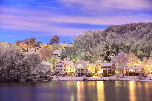 Snow-covered houses along a serene lake, illuminated by soft lights under a twilight sky.