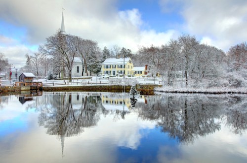 A serene winter scene with a snowy landscape, a church steeple, and reflections in a calm lake.