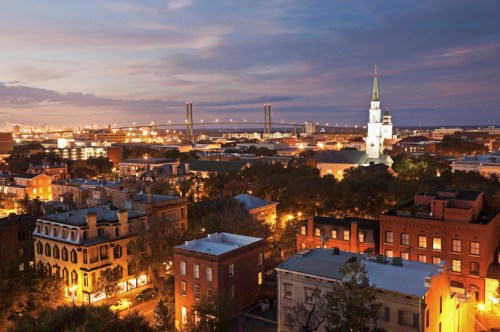 A city skyline at dusk, featuring buildings, a church steeple, and a bridge illuminated by city lights.