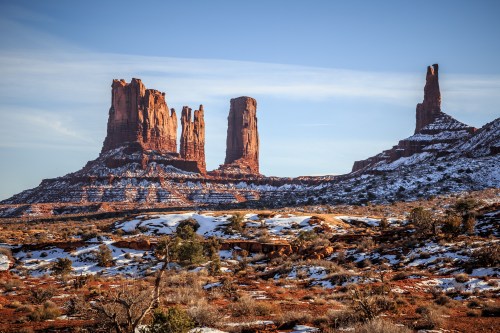 Majestic red rock formations rise against a clear sky, with patches of snow on the ground in a desert landscape.