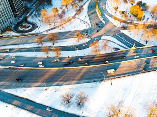 Aerial view of a snowy road with cars, surrounded by trees and buildings, casting long shadows in the winter sunlight.