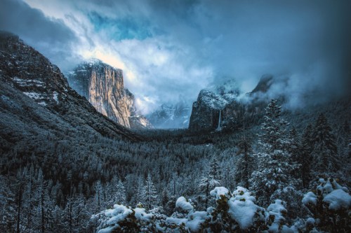 A snowy landscape in Yosemite National Park, featuring towering cliffs and misty mountains under a cloudy sky.