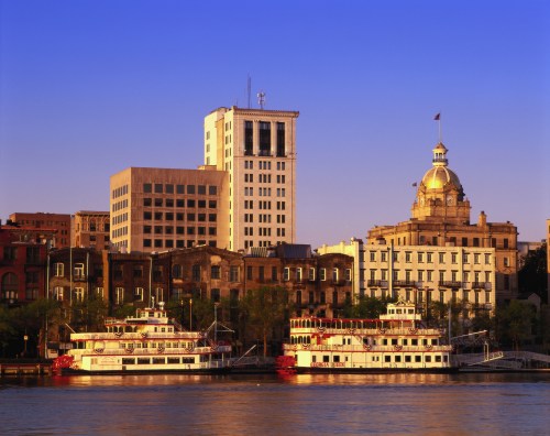 Two historic riverboats docked along a waterfront, with city buildings and a clear blue sky in the background.