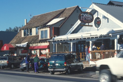 A cozy café with a wooden deck, people outside, and cars parked nearby, set in a snowy mountain town.