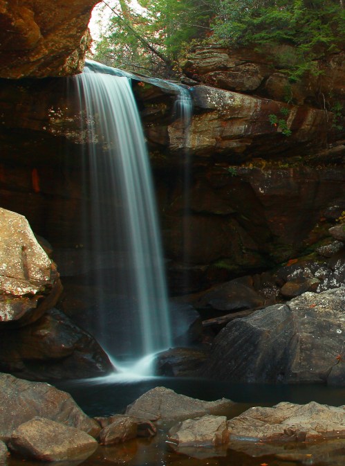 A serene waterfall cascades over rocks into a calm pool, surrounded by lush greenery and rugged stone formations.